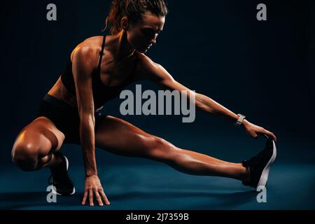 Sportler macht Dehnübungen auf schwarzem Hintergrund. Weibliche Läuferin dehnt die Beinmuskeln, indem sie seine Schuhe berührt, während sie im Studio sitzt. Stockfoto