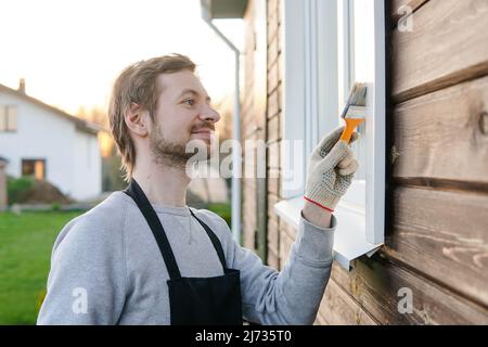Junger lächelnder Mann im Schürze malte Fenster seines Hauses mit Pinsel. Stockfoto
