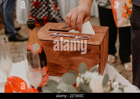 Eine Männerhand legt einen Umschlag mit Geld in eine festliche Holzkiste für Hochzeitsgeschenke an das Brautpaar. Slawische ukrainische russische Traditionen Stockfoto