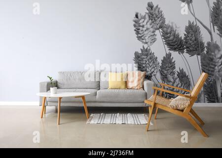 Interieur des stilvollen Wohnzimmers mit modernem Sessel, Tisch und Sofa in der Nähe der hellen Wand mit bedruckten Zweigen Stockfoto