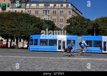 Eine Straßenbahn und zwei Personen, die im Sommer auf Radwegen am Strandvägen, am Ufer von Ostermalm, Stockholm, Schweden, fahren. Stockfoto