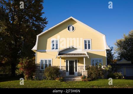 Gelbes zweistöckiges Haus mit Stein- und Holzplanken im Scheunenstil mit landschaftlich gestaltetem Vorgarten im Herbst. Stockfoto