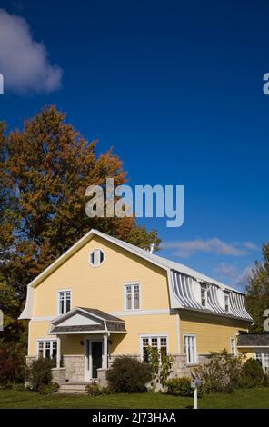 Gelbes zweistöckiges Haus mit Stein- und Holzplanken im Scheunenstil mit landschaftlich gestaltetem Vorgarten im Herbst. Stockfoto