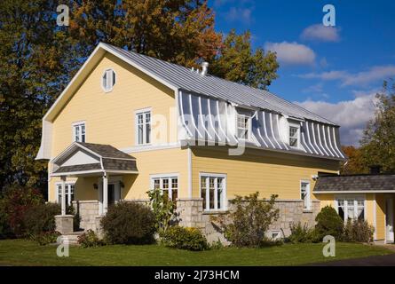 Gelbes zweistöckiges Haus mit Stein- und Holzplanken im Scheunenstil mit landschaftlich gestaltetem Vorgarten im Herbst. Stockfoto