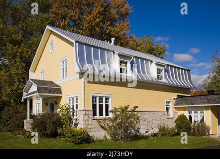 Gelbes zweistöckiges Haus mit Stein- und Holzplanken im Scheunenstil mit landschaftlich gestaltetem Vorgarten im Herbst. Stockfoto