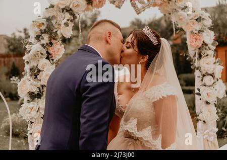 Braut und Bräutigam in einem Hochzeitskleid und einem langen Schleier bei einer Hochzeitszeremonie auf dem Hintergrund eines Blumenbogens lachen küssen jubelnd. Slawisch Stockfoto