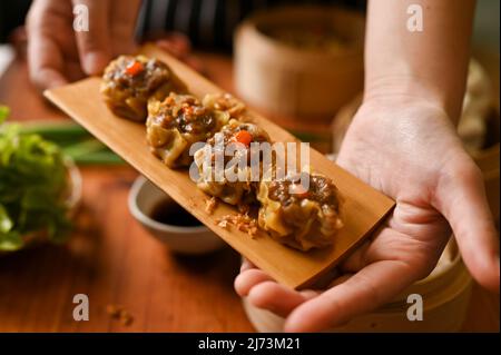 Weibliche Hände, die einen Teller gedünsteter Schweineknödel über dem Esstisch halten. Traditionelles chinesisches Essen Stockfoto