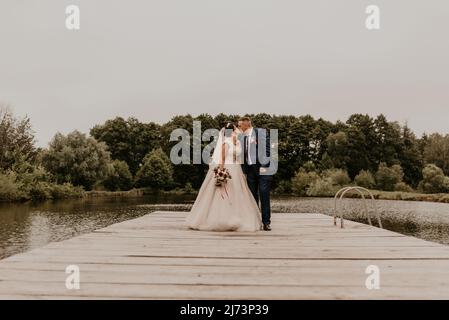 Blonde europäische kaukasische junge Mann Bräutigam in blauem Anzug und schwarzhaarige Frau Braut in weißem Hochzeitskleid mit langem Schleier und Tiara auf dem Kopf. Frisch Vermählt Stockfoto