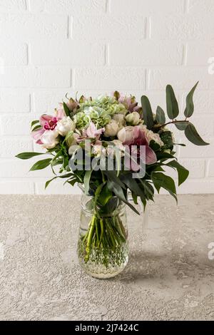 Ein schöner Strauß Gartenrosen in einer Glasvase auf einem grauen Betontisch an einer großen Ziegelwand. Das Konzept eines Geschenks, eine rhombtische Stimmung Stockfoto