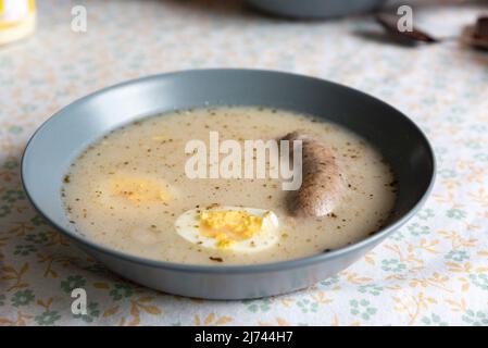 Ostersauer-Roggensuppe mit Weißwurst und Ei. Traditionelles polnisches Gericht zu Ostern. Saure Roggensuppe auf einem Teller und auf einer Tischdecke mit Mustern. Stockfoto