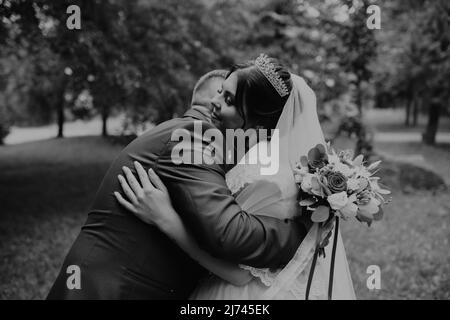 Blonde europäische kaukasischen jungen Mann Bräutigam in blauem Anzug und schwarzhaarige Frau Braut in weißem Hochzeitskleid mit langem Schleier und Tiara auf dem Kopf. Zuerst Stockfoto