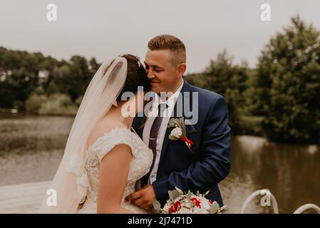 Blonde europäische kaukasische junge Mann Bräutigam in blauem Anzug und schwarzhaarige Frau Braut in weißem Hochzeitskleid mit langem Schleier und Tiara auf dem Kopf. Frisch Vermählt Stockfoto