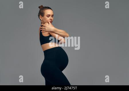 Cute schwanger Bauch isoliert auf grau. Seitenansicht einer jungen Schwangeren, die sich mit den Händen umarmt. Großer Bauch auf dem dritten Trimester der Schwangerschaft Stockfoto