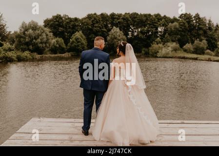 Blonde europäische kaukasische junge Mann Bräutigam in blauem Anzug und schwarzhaarige Frau Braut in weißem Hochzeitskleid mit langem Schleier und Tiara auf dem Kopf. Frisch Vermählt Stockfoto