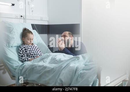 Junger trauriger verzweifelter Vater, der im Krankenhausbett für ein krankes kleines Mädchen unter Behandlung betet. Unwohl Kind ruht im Kinderklinikraum, während Pflege Eltern für ihre Gesundheit beten. Stockfoto