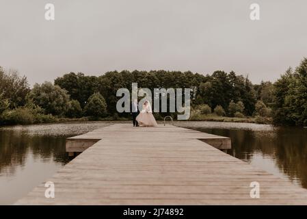 Blonde europäische kaukasische junge Mann Bräutigam in blauem Anzug und schwarzhaarige Frau Braut in weißem Hochzeitskleid mit langem Schleier und Tiara auf dem Kopf. Frisch Vermählt Stockfoto