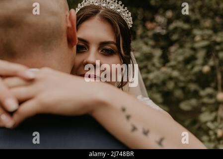 Holovchyntsi, Oblast Khmelnytska, Großbritannien raine - 07.12.2020: Braut im Schleier und eine Tiara auf dem Kopf umarmt den Bräutigam und lächelt auf dem Arm der Frau Stockfoto