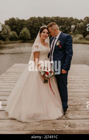 Blonde europäische kaukasische junge Mann Bräutigam in blauem Anzug und schwarzhaarige Frau Braut in weißem Hochzeitskleid mit langem Schleier und Tiara auf dem Kopf. Frisch Vermählt Stockfoto