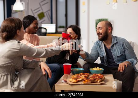 Eine vielfältige Gruppe von Arbeitskollegen, die bei einer Bürofeier mit Getränken und Snacks im Büro Gläser und Flaschen anklirren. Kollegen feiern mit einem alkoholischen Getränk und machen Prost Toast. Stockfoto