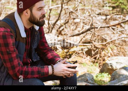 Junger bärtiger Tourist, der auf einem Felsen sitzt, ein zwei-Wege-Radio hält und nachdenklich in die Ferne blickt Stockfoto