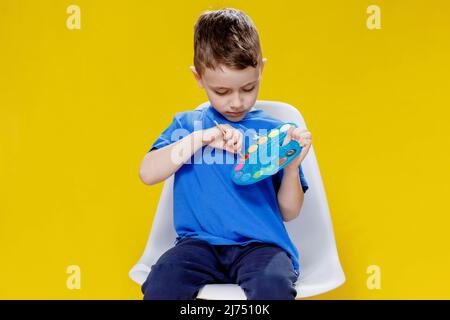 Kleiner netter Vorschuljunge, der eine Palette von Farben und einen Pinsel auf gelbem Hintergrund intensiv hält. Kreatives Kind. Stockfoto