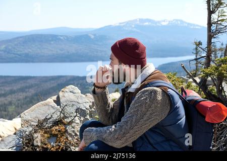 Wandern in den Bergen. Junger bärtiger Mann mit Rucksack, der auf der Bergspitze ruht, raucht und den malerischen Blick auf die wilde Natur genießt Stockfoto