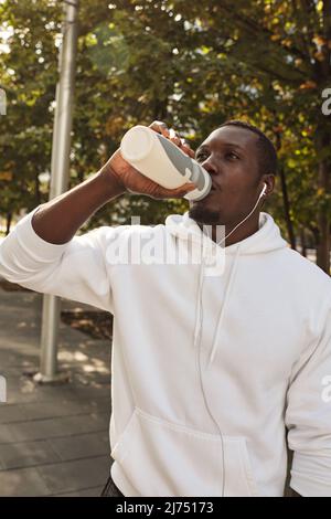 Junger durstiger, sportlicher schwarzer Mann mit weißem Hoodie und Trinkwasser aus der Sportflasche nach langen Laufstrecken im Freien Stockfoto
