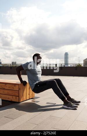 Der junge, athletische schwarze Mann in aktiver Trainingsbekleidung lehnt sich auf eine Holzbank und macht Liegestütze auf einem modernen Gewerbegebiet Stockfoto