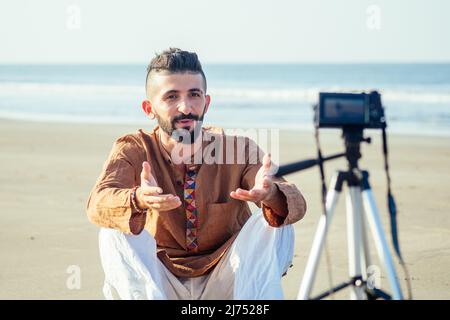 arabischer Mann sitzt auf Sand und spricht Stativ Video machen am Strand. Lifestyle-Content-Maker-Konzept für Reisevideografen Stockfoto