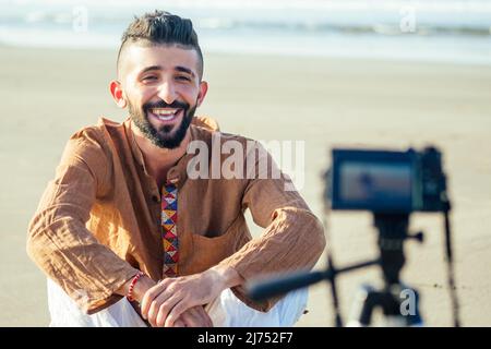 arabischer Mann sitzt auf Sand und spricht Stativ Video machen am Strand. Lifestyle-Content-Maker-Konzept für Reisevideografen Stockfoto