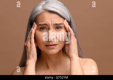 Die grauhaarige Asiatin mittleren Alters leidet unter starken Kopfschmerzen, blickt mit einem Anblick voller Schmerz und Verzweiflung auf die Kamera und massiert Schläfen in der Hoffnung zur Erleichterung Stockfoto