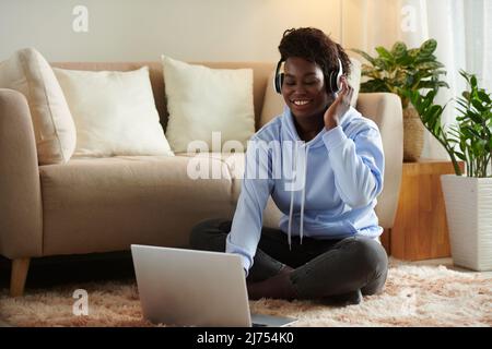 Fröhliche junge Frau mit Kopfhörern, die auf dem Boden sitzt und dem neuen Lied ihrer Lieblingsmusikband lauscht Stockfoto