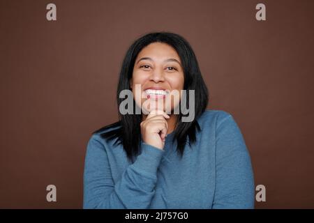 Portrait einer aufrichtigen jungen schwarzen Frau, die lacht und dabei das Kinn vor braunem Hintergrund berührt Stockfoto