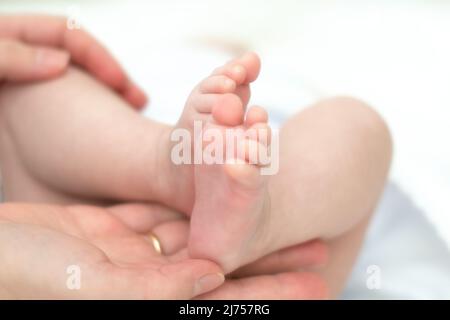 Die Hände der Mutter halten zärtlich und liebevoll die Beine eines kleinen Kindes, das seit der Geburt mehrere Monate alt ist. Stockfoto