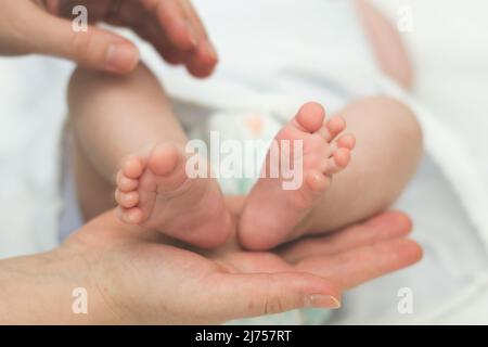 Die Hände der Mutter halten zärtlich und liebevoll die Beine eines kleinen Kindes, das seit der Geburt mehrere Monate alt ist. Stockfoto