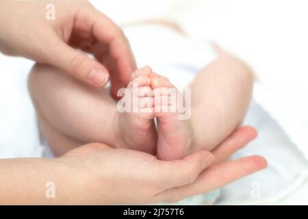 Die Hände der Mutter halten zärtlich und liebevoll die Beine eines kleinen Kindes, das seit der Geburt mehrere Monate alt ist. Stockfoto