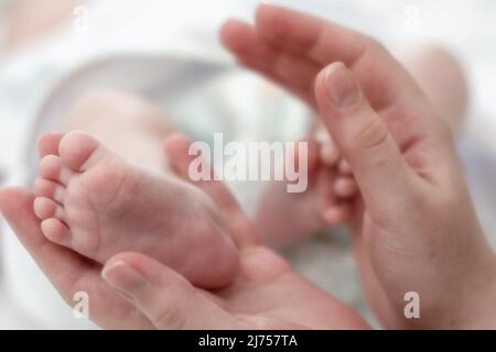 Die Hände der Mutter halten zärtlich und liebevoll die Beine eines kleinen Kindes, das seit der Geburt mehrere Monate alt ist. Stockfoto