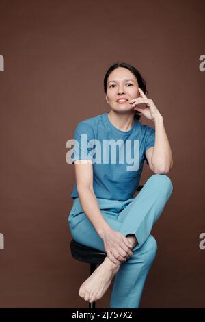 Porträt einer lächelnden attraktiven Dame mittleren Alters in blauem Outfit, die auf einem Stuhl sitzt und das Gesicht vor dunklem Hintergrund berührt Stockfoto