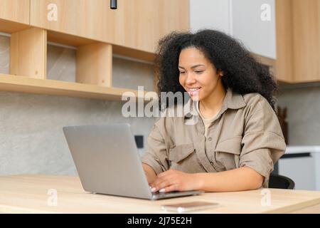 Glückliche und konzentrierte Geschäftsfrau in Freizeitkleidung schaut auf den Laptop-Bildschirm, löst Aufgaben, freiberufliche Frau arbeitet von zu Hause aus Stockfoto