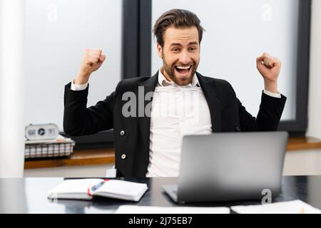 Glücklicher männlicher Büroangestellter in formeller Kleidung, der im Triumph die Fäuste hochhob, gute Nachrichten feierte, einen unglaublichen Deal erhielt, Geschäftsmann freut sich, im Büro zu sitzen und auf den Laptop-Bildschirm zu schauen. Gewinnen und Glück Stockfoto