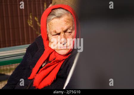 Defokussieren sie die ältere ukrainische Frau in einem roten Schal mit Krücken, die draußen sitzen. Frau 90 Jahre alt. Einzelperson. Nahaufnahme Porträt. Krank und heimatverbunden Stockfoto