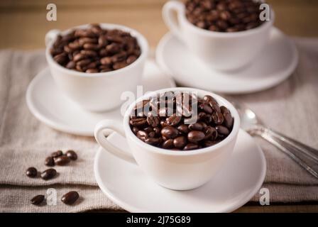 Drei weiße Kaffeetassen auf weißen Untertassen gefüllt mit Kaffeebohnen Stockfoto