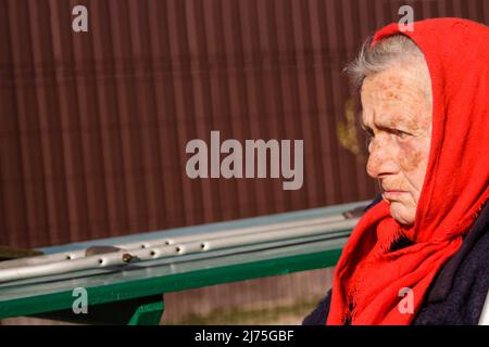 Defokussieren sie die ältere ukrainische Frau in einem roten Schal mit Krücken, die draußen sitzen. Frau 85 Jahre alt. Einzelperson. Nahaufnahme. Krank und heimatverbunden. Traurig Stockfoto