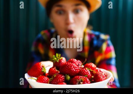 Unschärfe junge Frau in einem Hut und Hemd lächelt und zeigt eine weiße Schüssel mit Erdbeeren. Dunkelgrüner Stahlhintergrund. Sommeressen, Obst. Sommerzeit Stockfoto