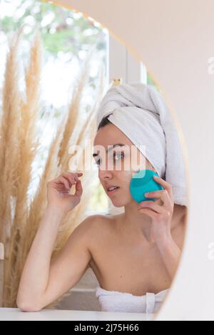 Junge Frau, die bei morgendlichen Hygieneverfahren im Bad die Gesichtshaut mit einem speziellen Ultraschallgerät säubert. Hautpflege- und Wellness-Konzept Stockfoto