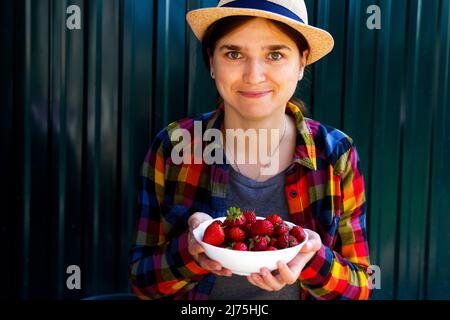 Unschärfe junge Frau in einem Hut und Hemd lächelt und zeigt eine Schüssel mit Erdbeeren. Dunkelgrüner Stahlhintergrund. Sommeressen, Obst. Sommerhintergrund Stockfoto