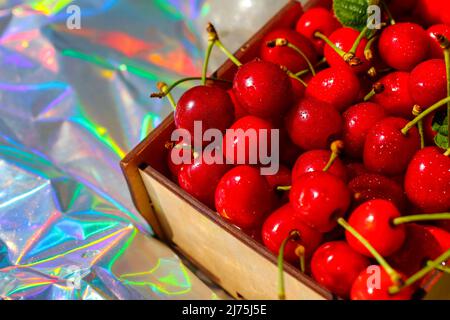 Unschärfe-Nahaufnahme-Box, Crape von dunkelroten, nassen Süßkirschen mit Schwanz und Blättern auf modernem Hologramm-Hintergrund. Früchte und Beeren. Ernte und Stockfoto