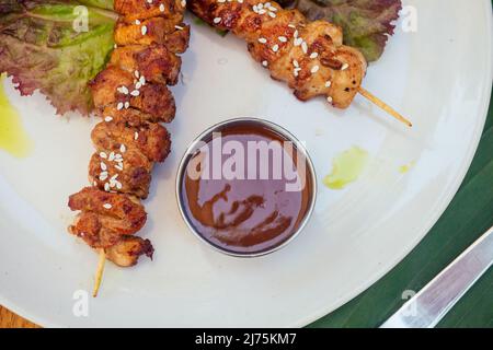 asiatischer Kebab im tropischen Stil auf dem Teller mit Bananen- und Salatblättern und Sesamsamen auf dem Holztisch Stockfoto
