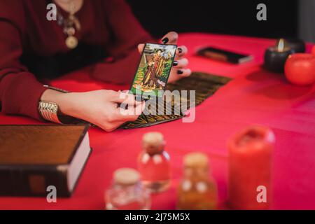 KIEW, UKRAINE - 23. FEBRUAR 2022: Beschnittene Ansicht des Wahrsagers, der Tarot-Karte in der Nähe des Buches hält, und verschwommene Gläser auf dem Tisch, isoliert auf Schwarz Stockfoto