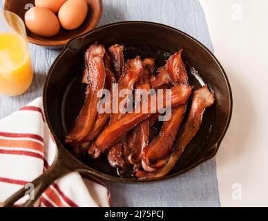 Knusprige Bacon-Streifen in einer gusseisernen Pfanne; ein Glas Orangensaft und eine Schüssel Eier Stockfoto